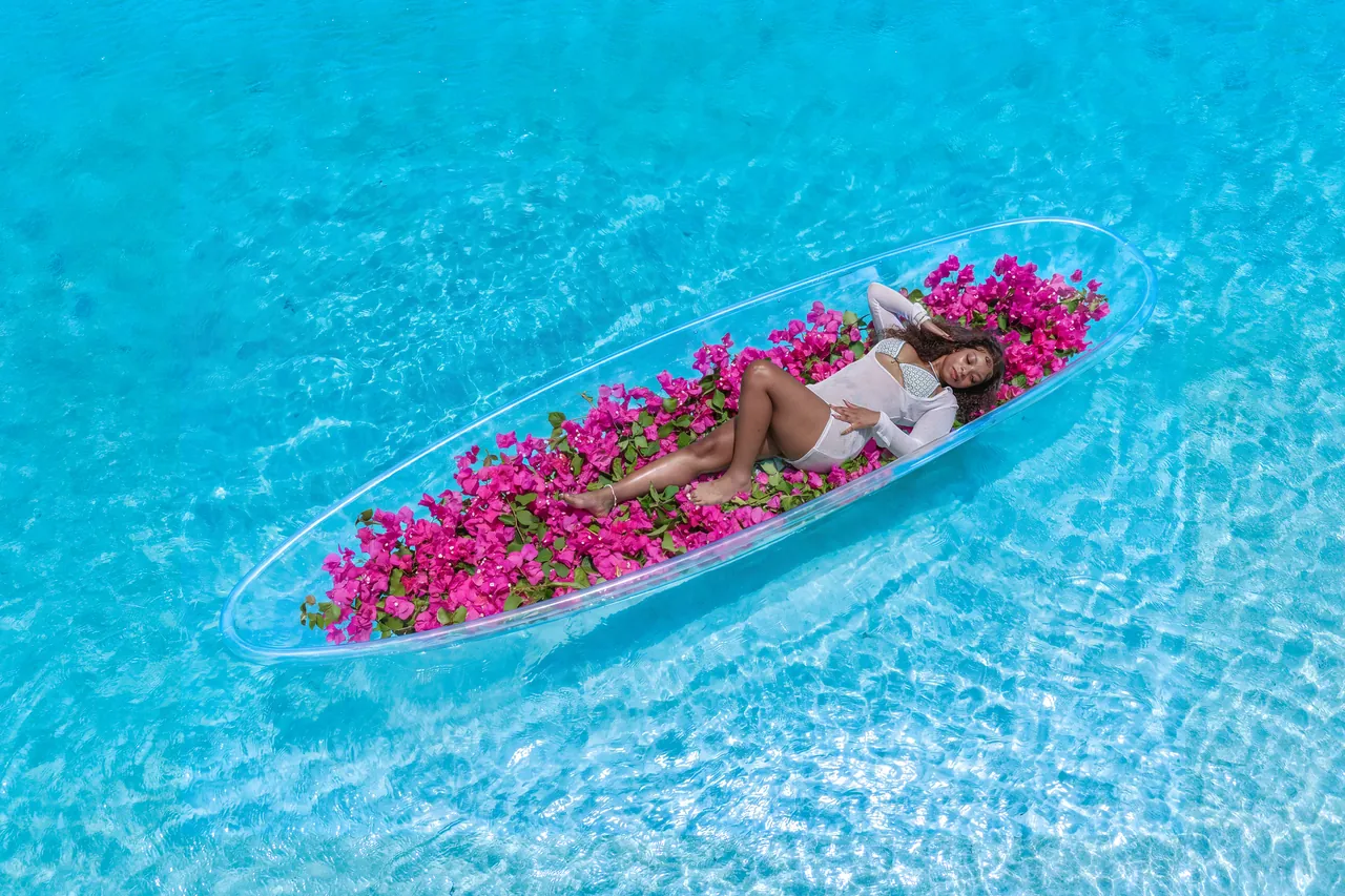 Guest reclining in a clear kayak filled with pink flowers over pale blue water